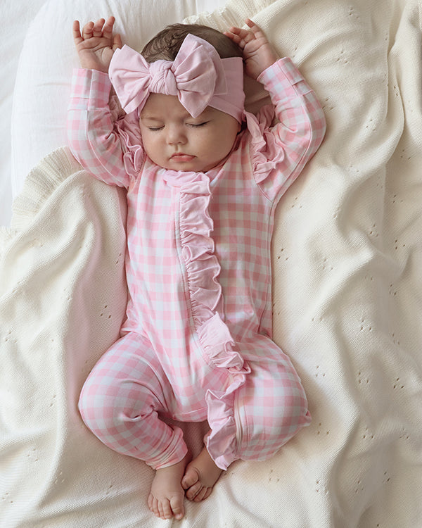 baby dressed in pink gingham frilly onesie with matching one size bow laying on white blanket
