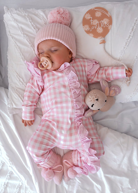 baby dressed in pink gingham frilly onesie with matching one size bow laying on white blanket