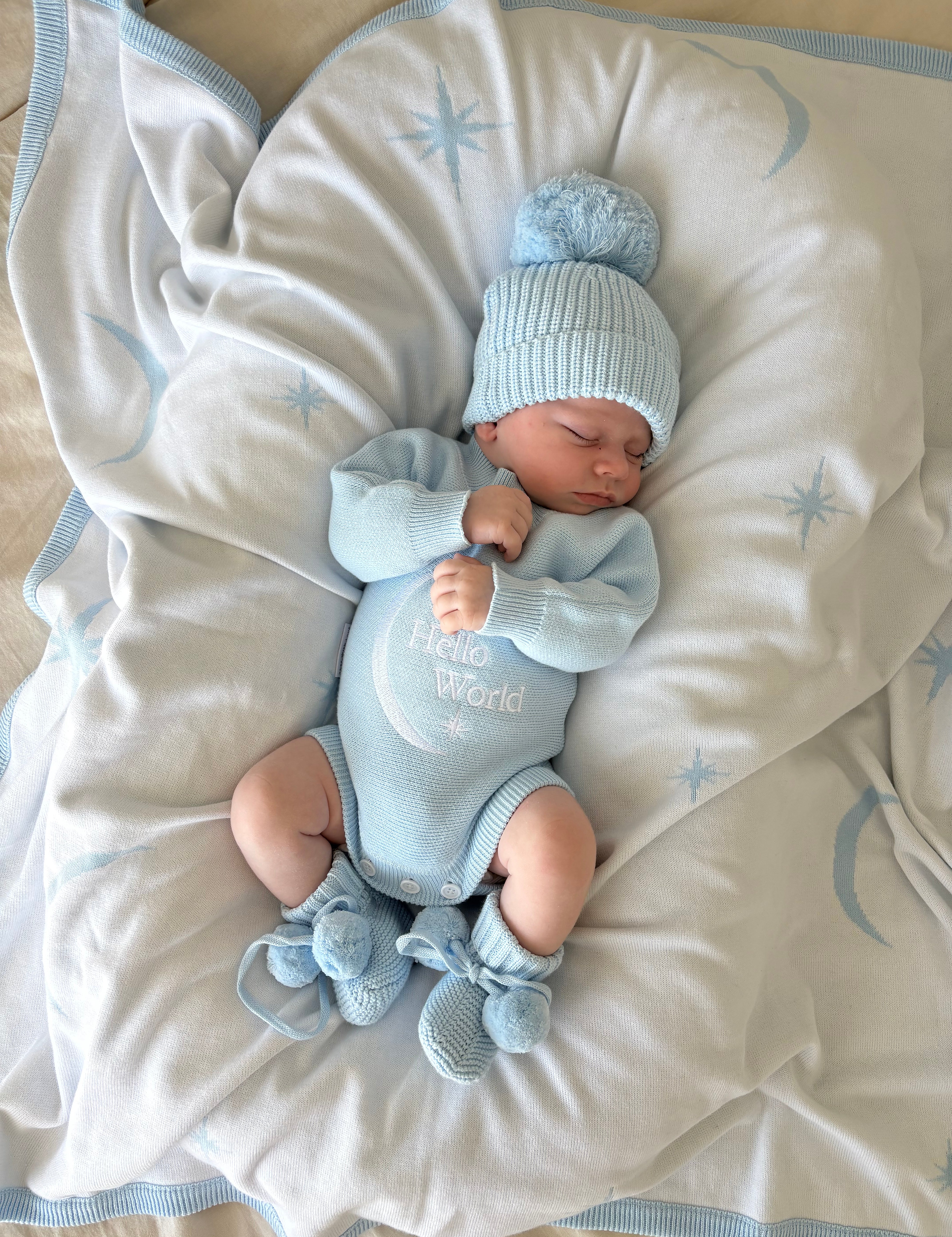 Newborn baby in baby blue hello world announcement romper and matching beanie and booties lying on a soft baby blue blanket with star and moon pattern with star patterns