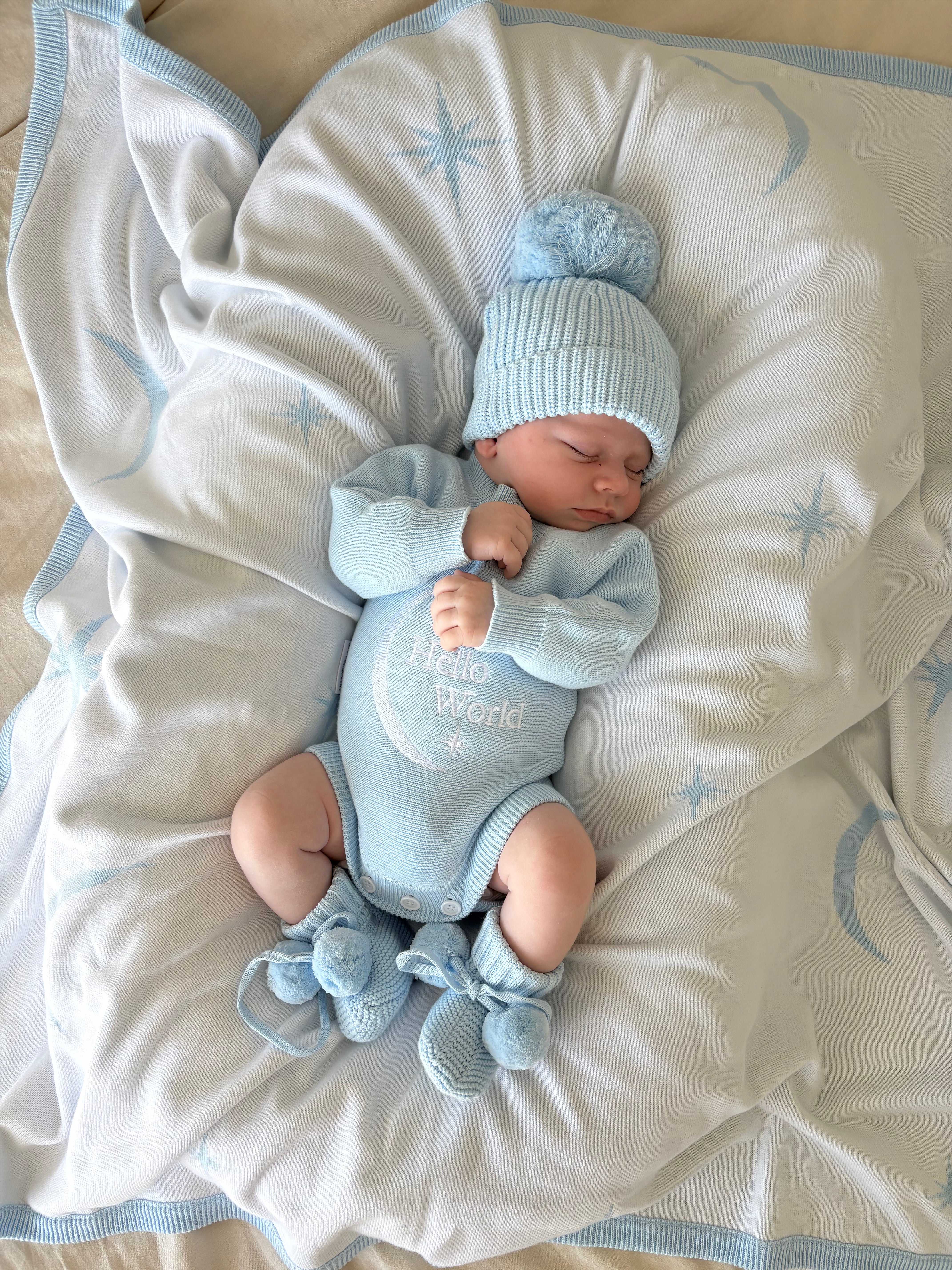 Newborn baby in baby blue hello world announcement romper and matching beanie and booties lying on a soft baby blue blanket with star and moon pattern with star patterns