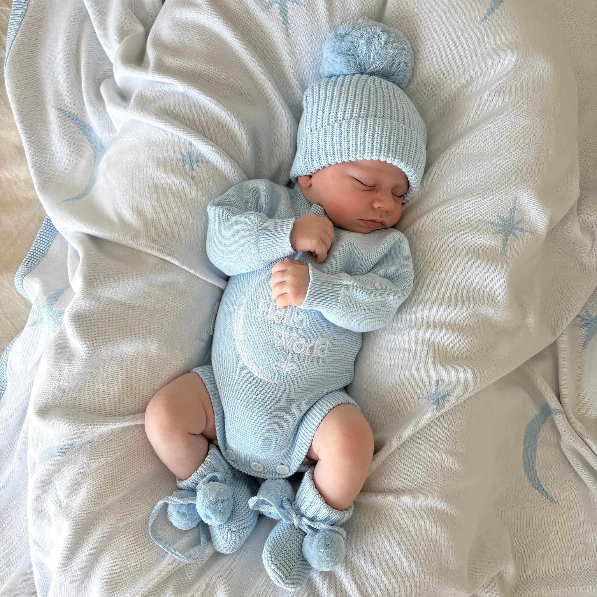 Newborn baby in baby blue hello world announcement romper and matching beanie and booties lying on a soft baby blue blanket with star and moon pattern with star patterns