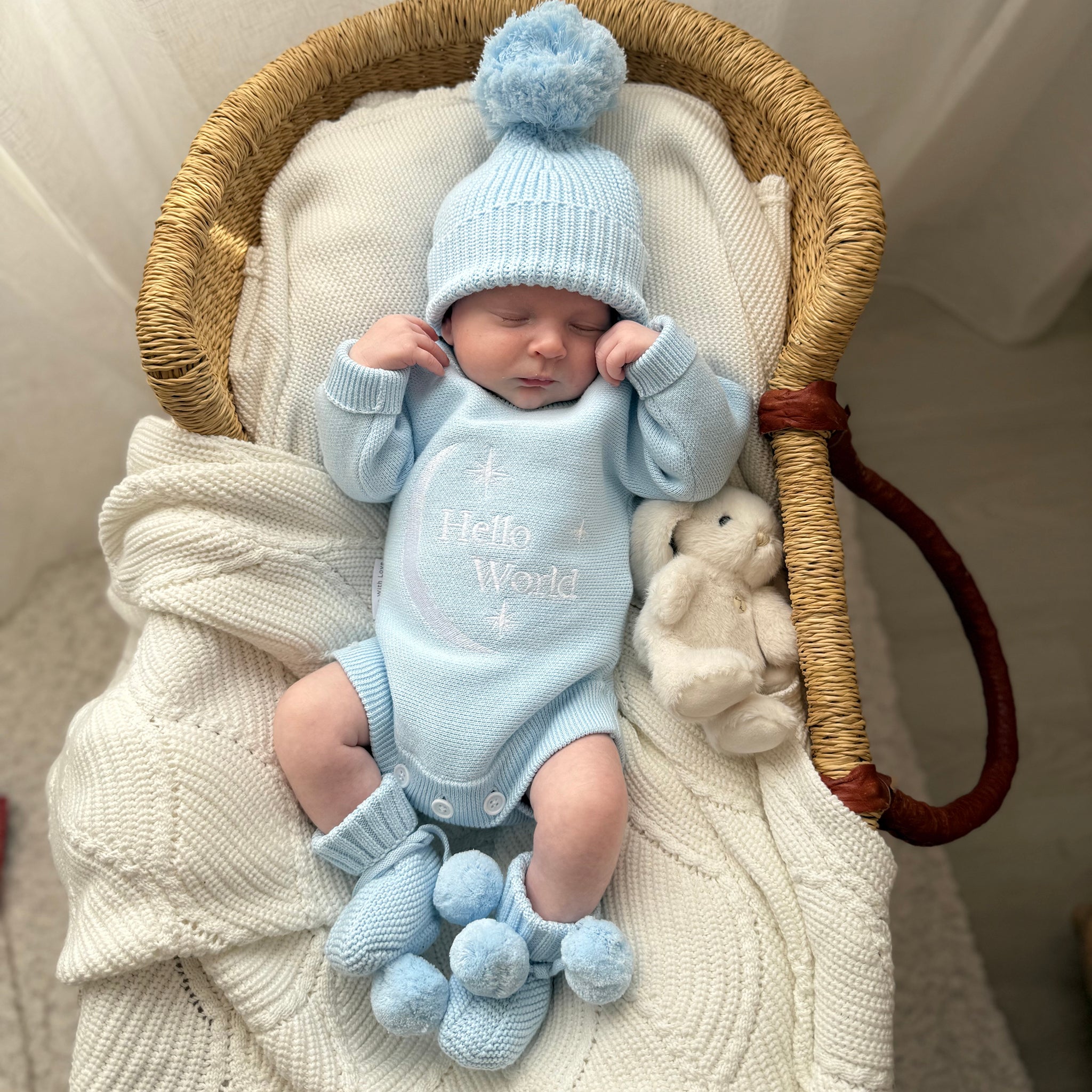 Newborn baby in a baby blue hello world outfit and beanie and booties with a teddy bear, lying in a woven basket.