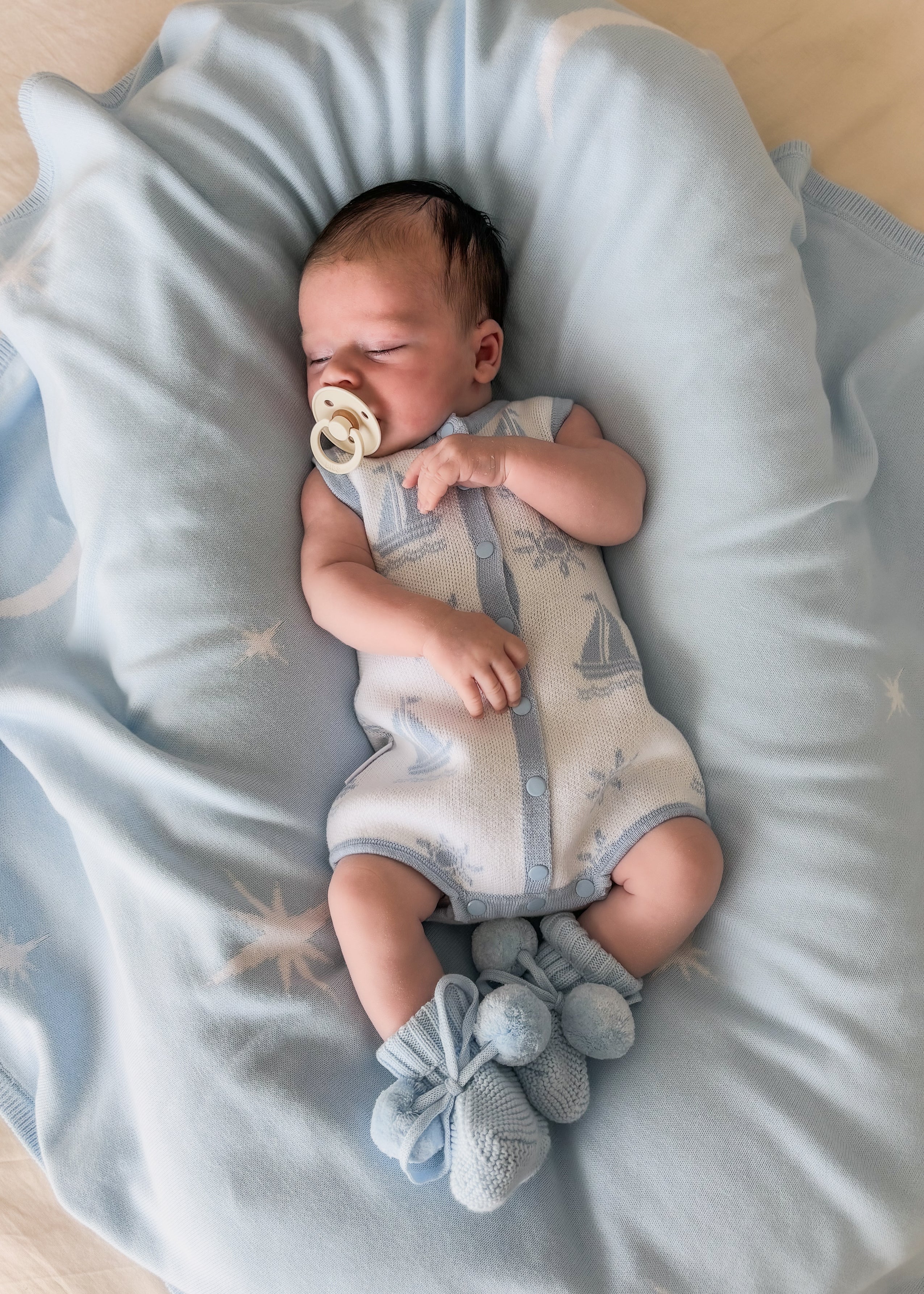 baby dressed in the coastal bubble romper laying on baby blue blanket wear baby blue booties and dummy in mouth