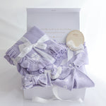 A lavender baby gift set arranged in a white box, including a ruffled blanket with white floral print, a matching bonnet with a bow, a pair of booties, and a small wooden tag that reads 'SHE'S'here 