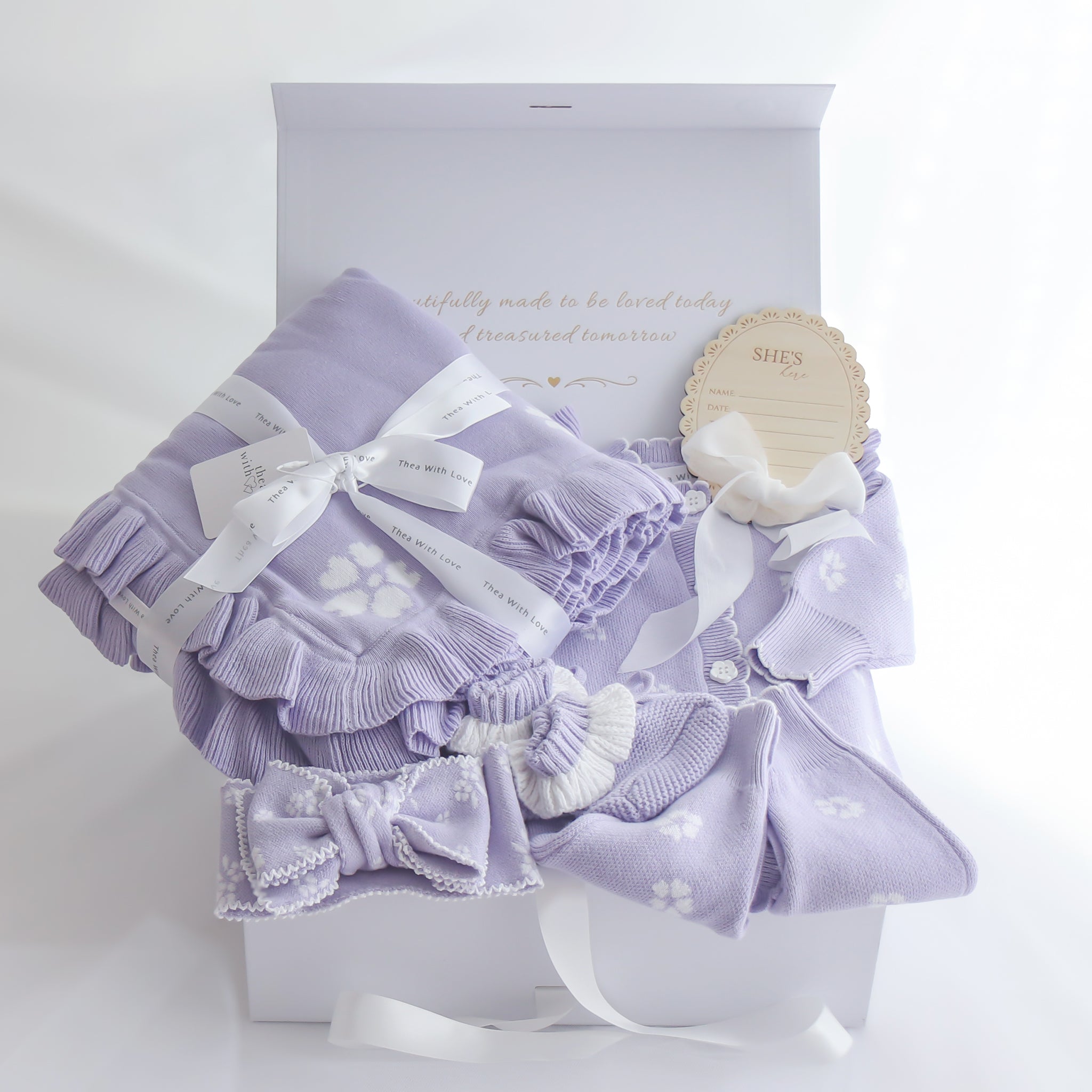 A lavender baby gift set arranged in a white box, including a ruffled blanket with white floral print, a matching bonnet with a bow, a pair of booties, and a small wooden tag that reads 'SHE'S'here 