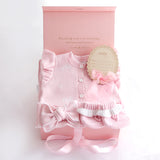 A pink baby gift set featuring a long-sleeve knit romper with ruffles and buttons, a matching bonnet with a bow, a round decorative tag, and an open pink gift box on a white background.