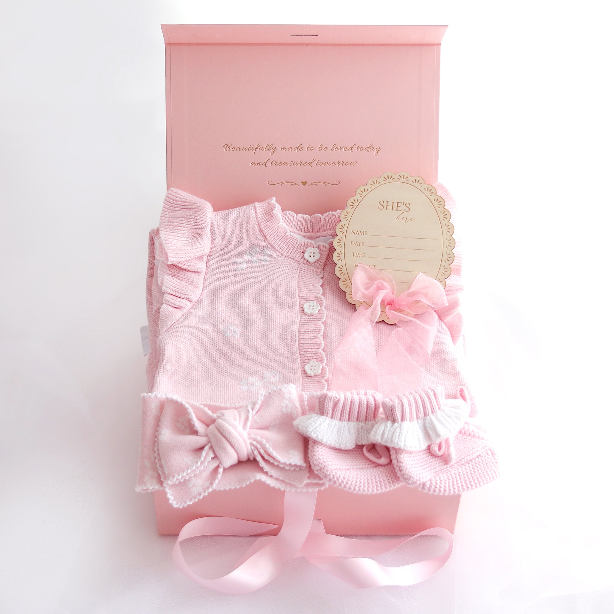 A pink baby gift set featuring a long-sleeve knit romper with ruffles and buttons, a matching bonnet with a bow, a round decorative tag, and an open pink gift box on a white background.