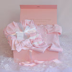 A pink baby gift set arranged inside an open pink box on a lace surface. The set includes a long-sleeve button-front baby cardigan, folded socks, a bow, and ribbons, with a small tag reading 'Baby with Love'.