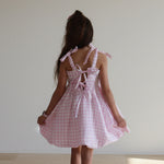 Person wearing a pink checkered dress standing in a room with a white wall and tiled floor. back of dress showing 2 bows in the middle of back and 2 bows on shoulder

