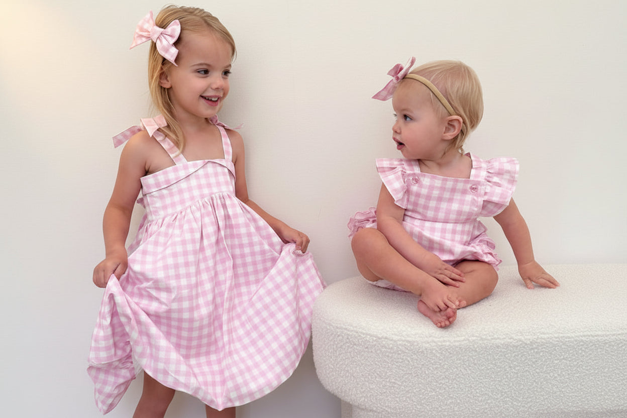 Two young girls in matching pink checkered dresses standing on a white surface.