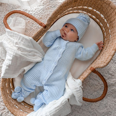 Baby in a blue knitted outfit and hat lying in a wicker basket.