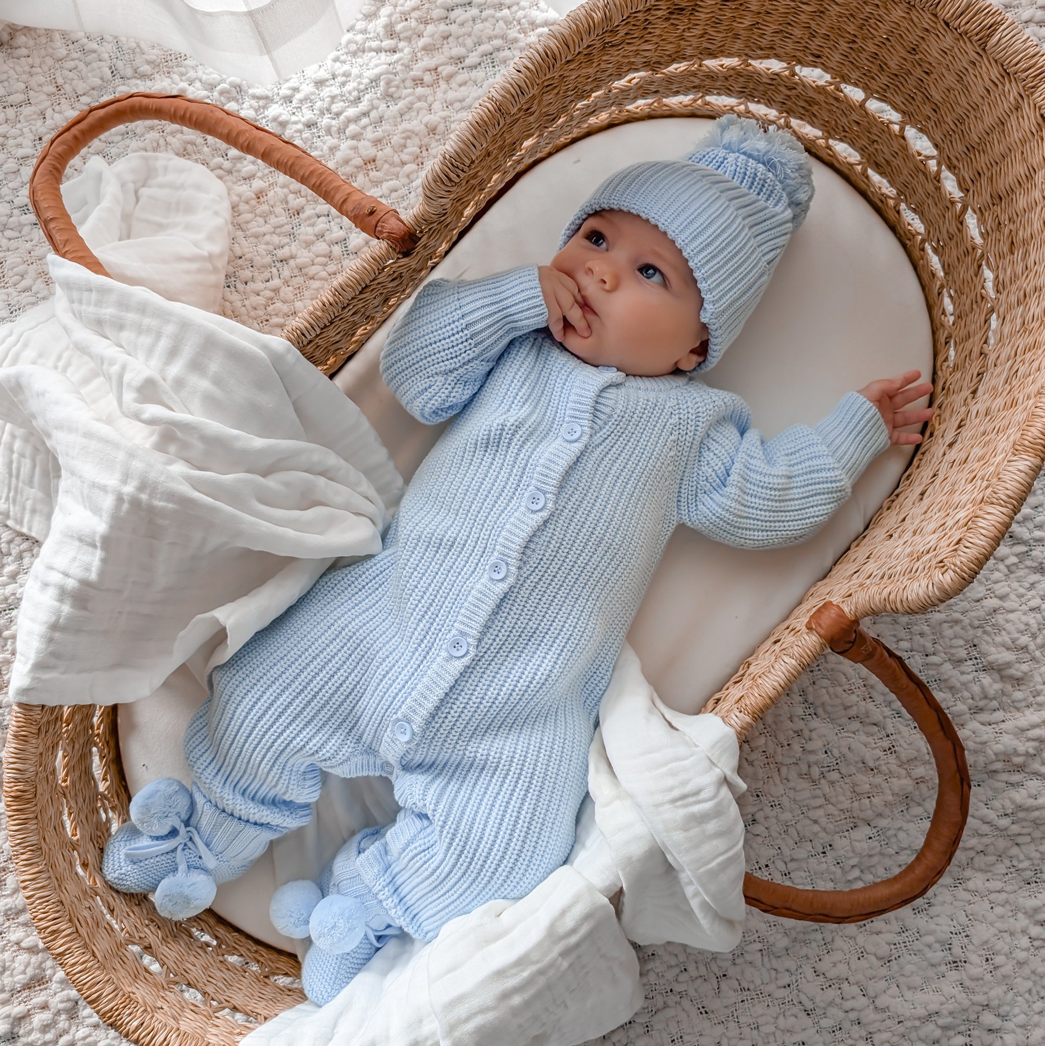 Baby in a blue knitted outfit and hat lying in a wicker basket.