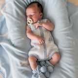 baby wearing coastal bubble romper with baby blue booties with pom poms laying on a baby blue blanket dummy in mouth