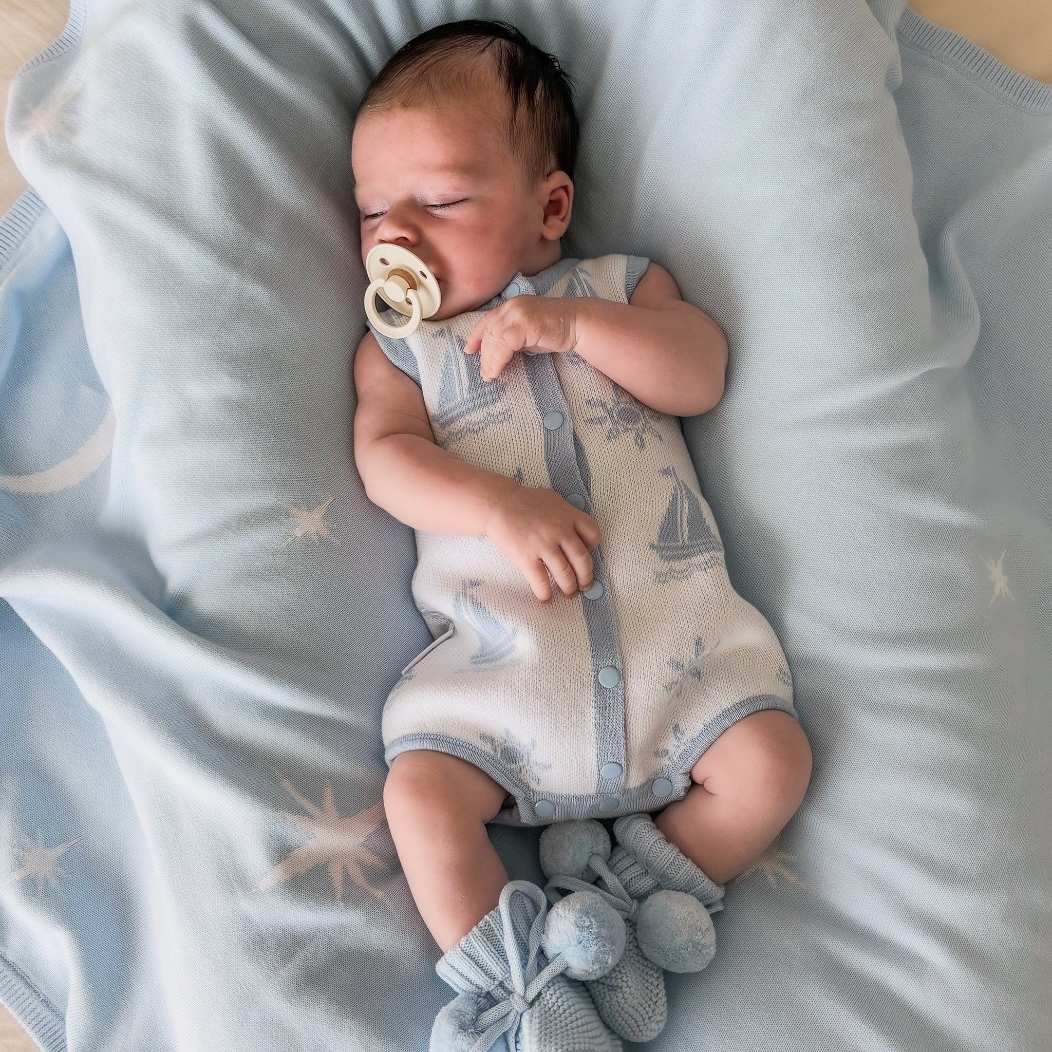 baby wearing coastal bubble romper with baby blue booties with pom poms laying on a baby blue blanket dummy in mouth