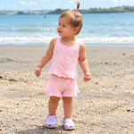 A toddler stands on a sandy beach wearing a light pink crochet knit two-piece outfit with a ruffled hem and front buttons, paired with lavender. The background shows the blue ocean and distant green shoreline.