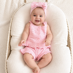 A smiling baby wearing a pink knit sleeveless romper with ruffle trim and a matching pink bow headband, posed on a cream muslin bassinet cushion.