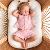 A sleeping baby girl wearing a pink knit two-piece outfit with a ruffled hem, lying on a white muslin bassinet cushion inside a wicker bassinet.