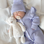 Newborn baby in a purple knit outfit with a matching beanie and pacifier, holding a plush toy on a white blanket.