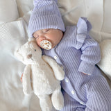 Newborn baby in a purple knit outfit with a matching beanie and pacifier, holding a plush toy on a white blanket.