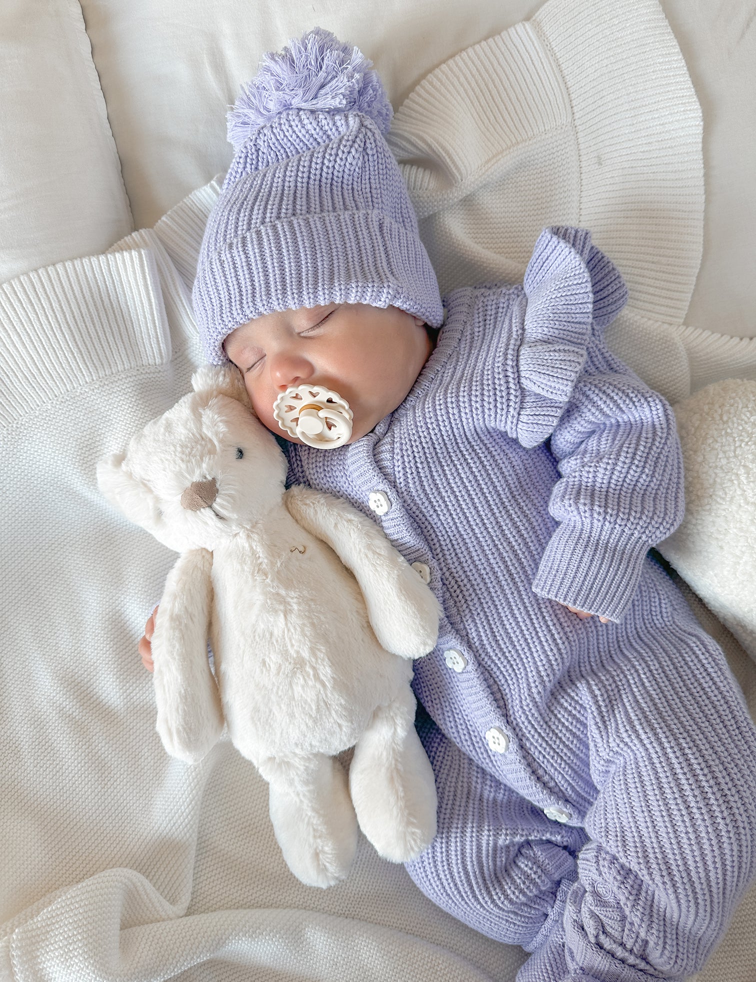 Newborn baby in a purple knit outfit with a matching beanie and pacifier, holding a plush toy on a white blanket.