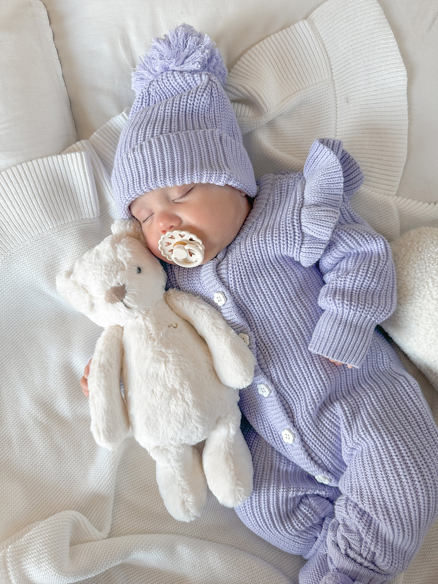 Newborn baby in a purple knit outfit with a matching beanie and pacifier, holding a plush toy on a white blanket.