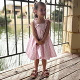 Young girl in a pink gingham dress standing on a wooden deck by a railing.