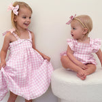 Two young girls in matching pink checkered dresses standing on a white surface.