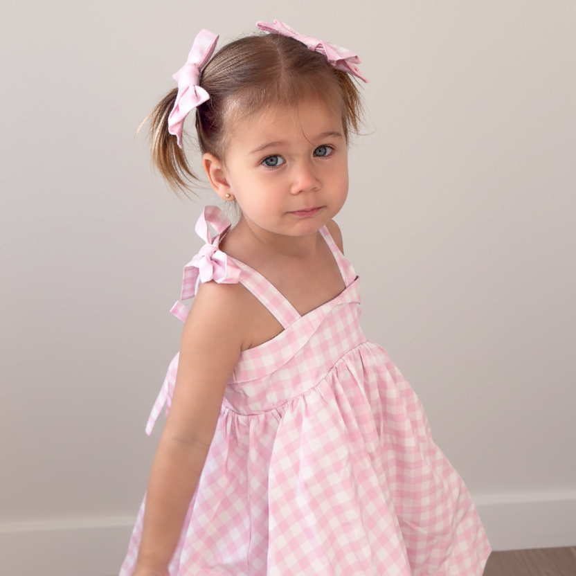 Young girl wearing a pink gingham dress with bow straps against a plain background