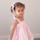 Young girl wearing a pink gingham dress with bow straps against a plain background