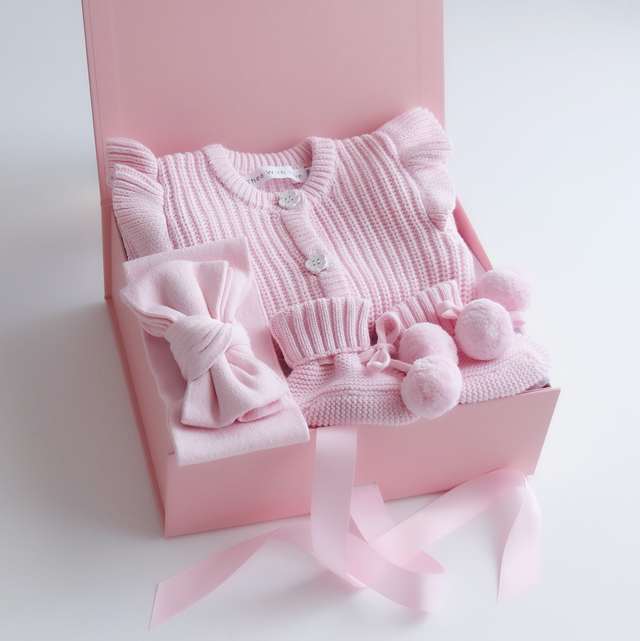 Pink baby romper outfit with bow and pom-pom booties in a pink box with pink satin ribbon on a white background