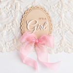 A wooden oval sign with the words 'IT'S A Girl' in white lettering, tied with a pink ribbon, displayed on a white lace background.