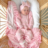 A sleeping baby in a pink knit onesie and matching bow headband, wrapped in a pink and white blanket, lying in a woven bassinet.