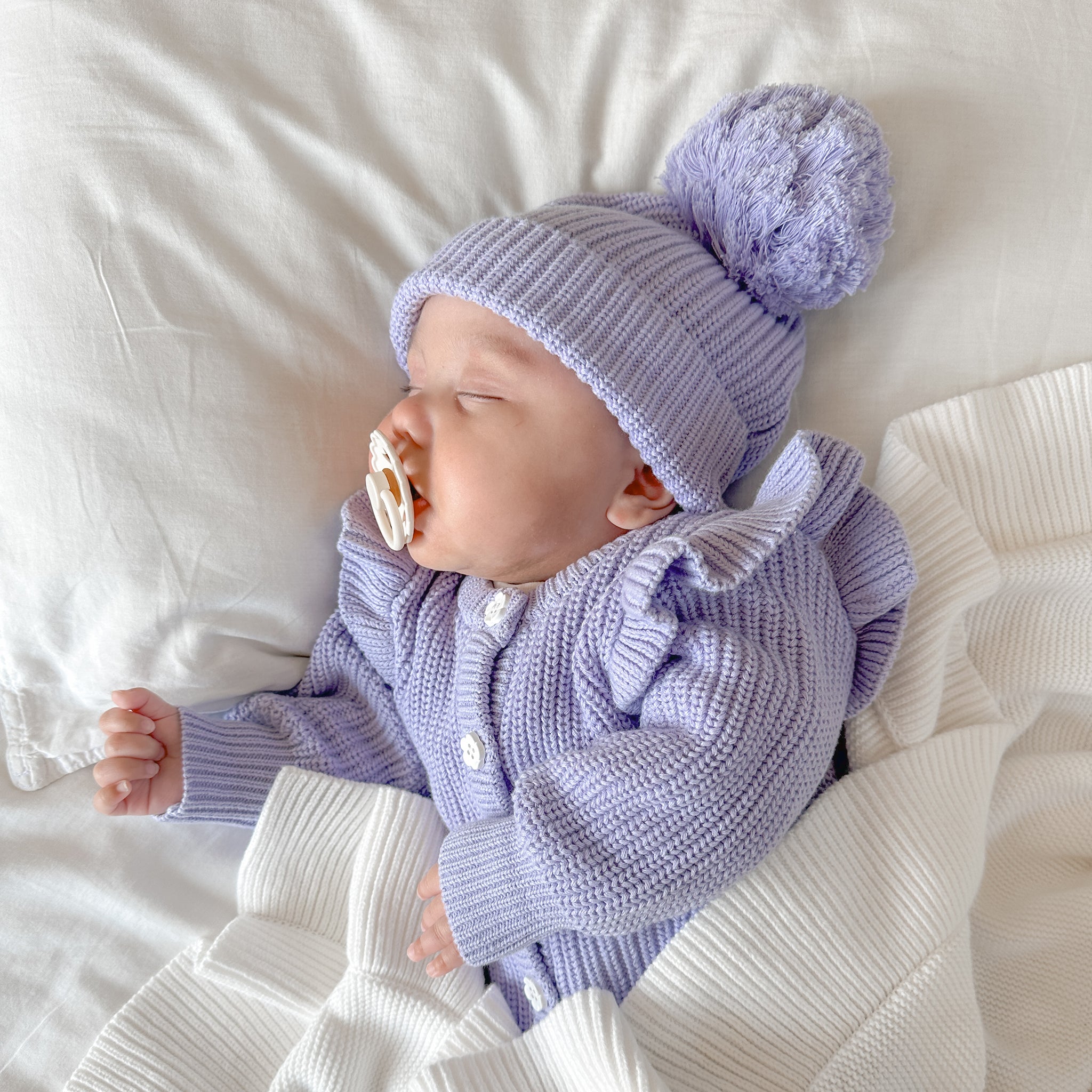 Baby wearing a purple knit outfit with a matching beanie on a white blanket