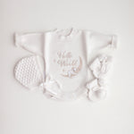 A flat lay of a white baby romper with 'Hello World' text and a koala illustration, accompanied by a bonnet and booties on a white background.