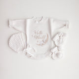 A flat lay of a white baby romper with 'Hello World' text and a koala illustration, accompanied by a bonnet and booties on a white background.