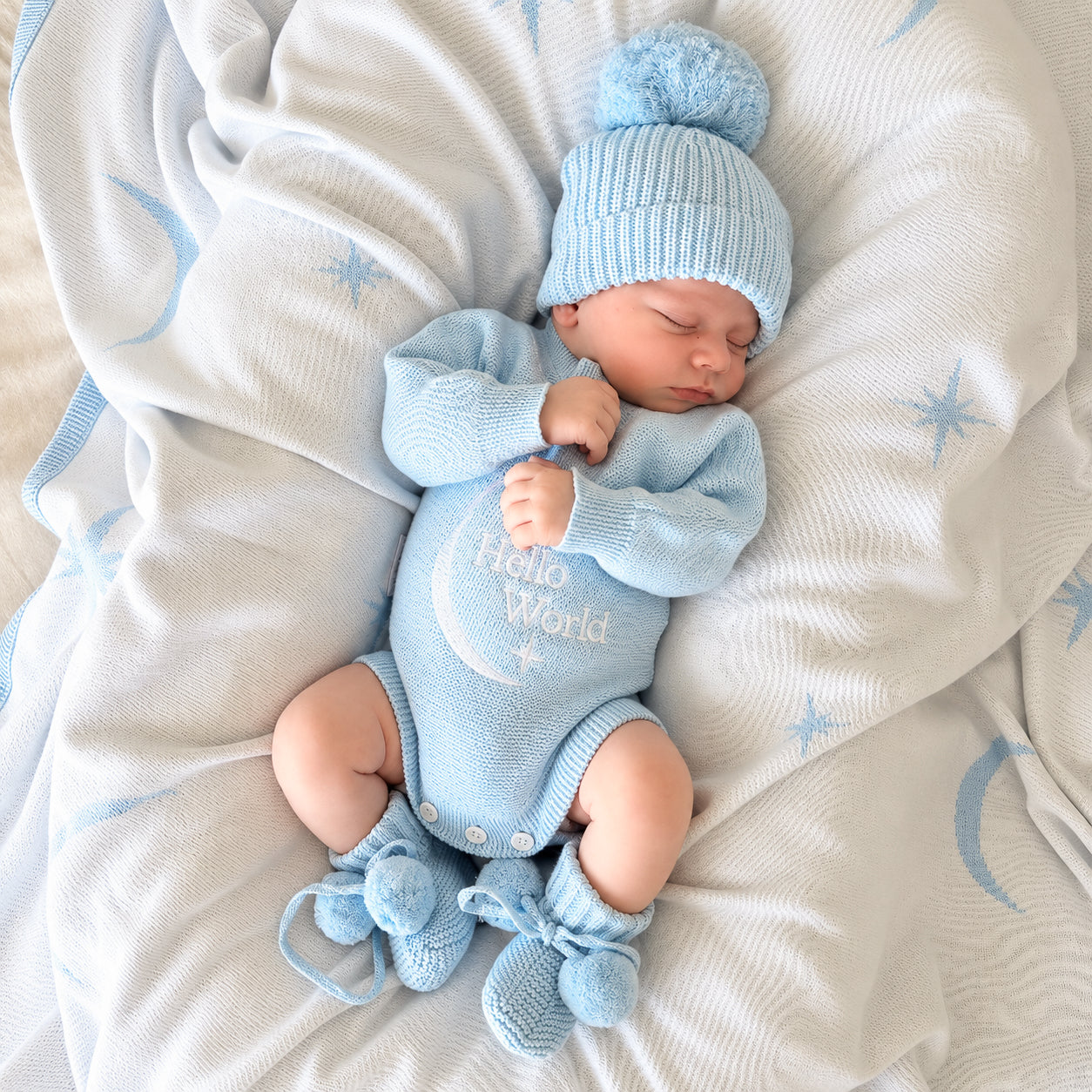 A sleeping newborn baby wearing a light blue knit onesie with 'Hello World' and a crescent moon design, along with a matching blue pom-pom beanie and booties, lying on a white blanket with blue star and moon patterns.

Details
