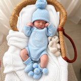 A sleeping newborn baby wearing a light blue knit bodysuit with 'Hello World' embroidered on the chest, a matching blue knit hat with a pom-pom, and blue booties, resting inside a woven bassinet with a white blanket and a small stuffed bunny.
