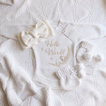 A baby gift set laid flat on a textured white knit blanket, including a white long-sleeve bodysuit with 'Hello World' and a moon-and-baby embroidery, a matching bow headband, knit booties with pom-poms, 