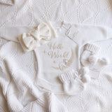 A baby gift set laid flat on a textured white knit blanket, including a white long-sleeve bodysuit with 'Hello World' and a moon-and-baby embroidery, a matching bow headband, knit booties with pom-poms, 
