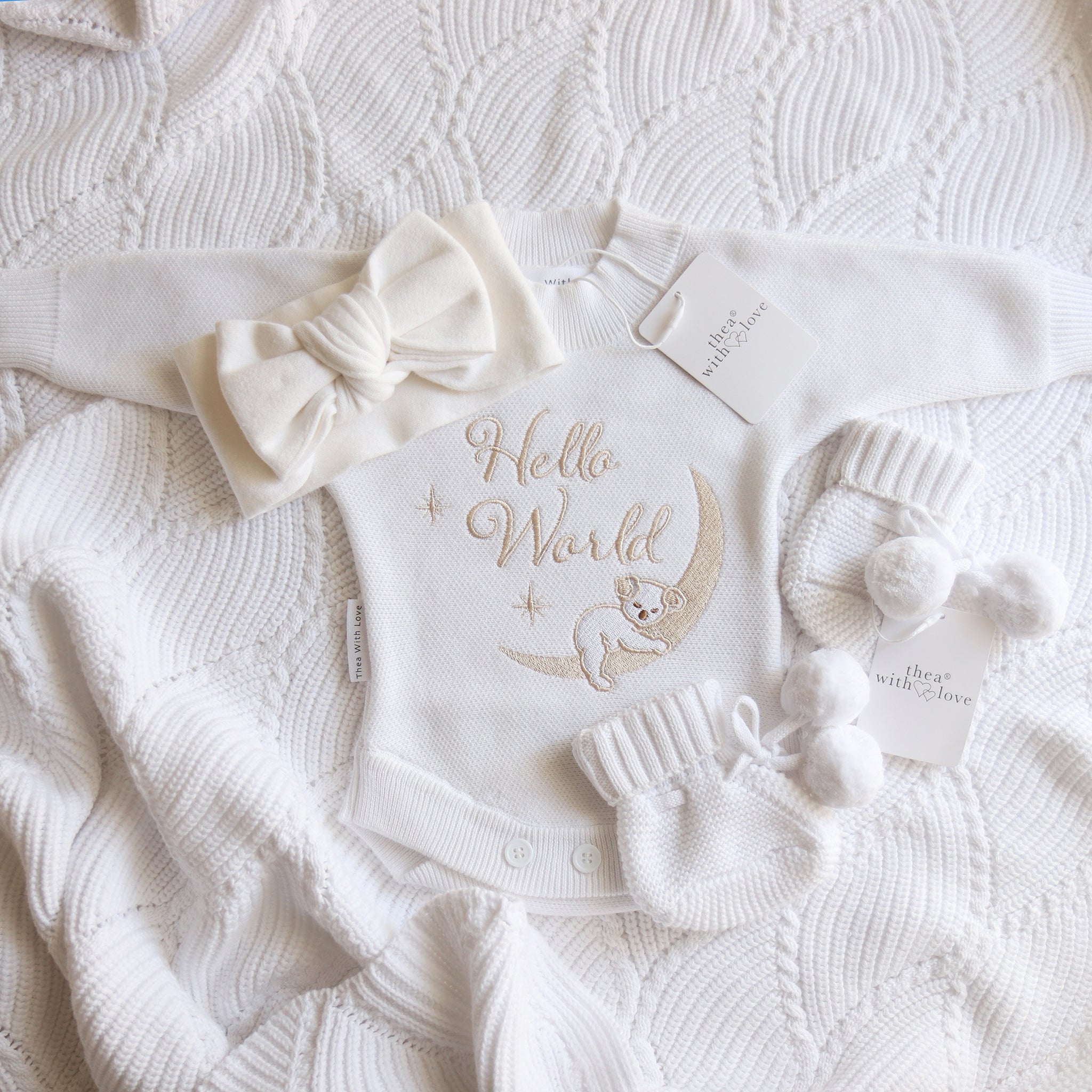 A baby gift set laid flat on a textured white knit blanket, including a white long-sleeve bodysuit with 'Hello World' and a moon-and-baby embroidery, a matching bow headband, knit booties with pom-poms, 