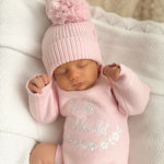A sleeping newborn baby wearing a light pink knit bonnet with a fluffy pom-pom and a matching pink long-sleeve bodysuit with 'Hello World' embroidered on the front, lying on a white textured blanket.