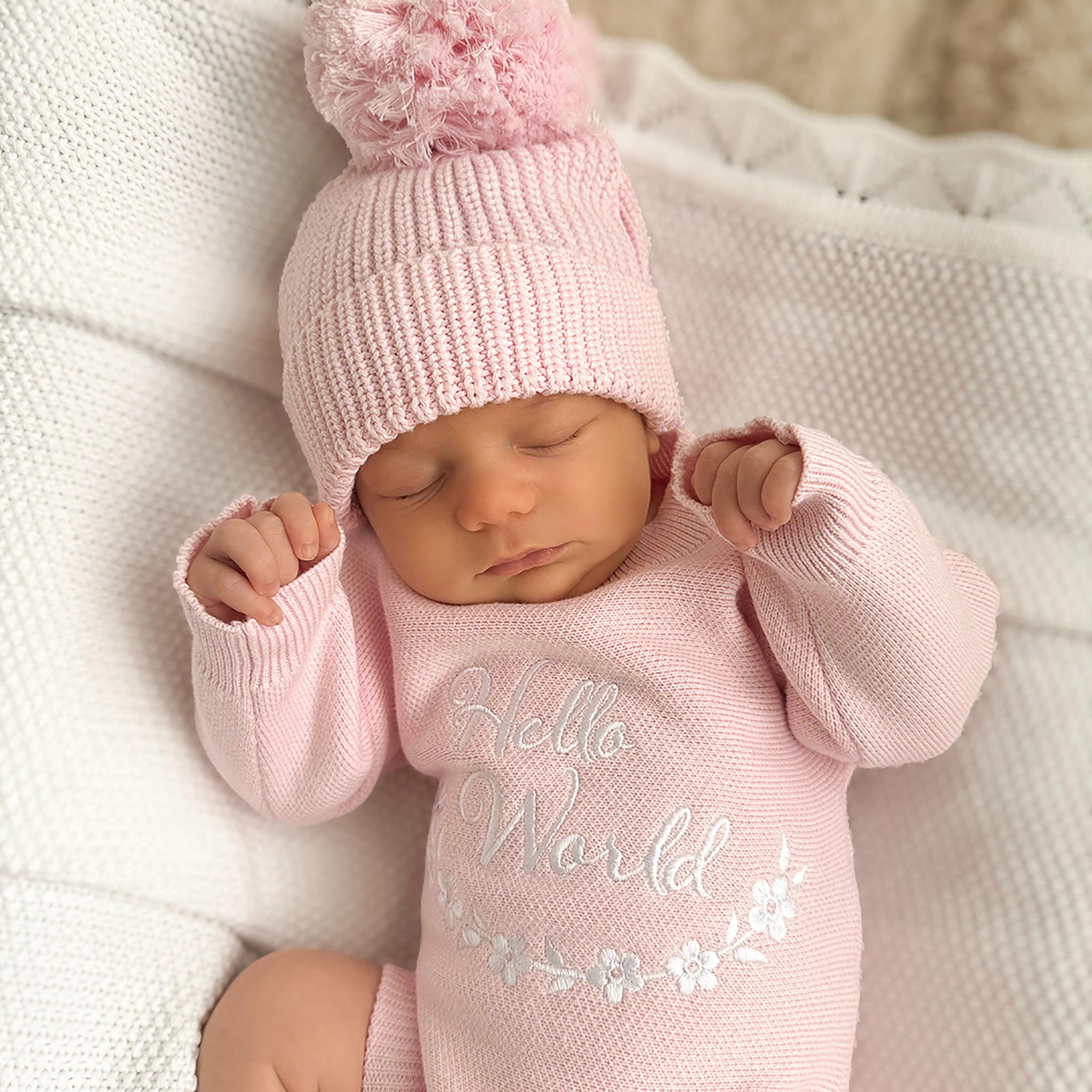 A sleeping newborn baby wearing a light pink knit bonnet with a fluffy pom-pom and a matching pink long-sleeve bodysuit with 'Hello World' embroidered on the front, lying on a white textured blanket.