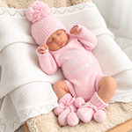A sleeping newborn baby wearing a pink knit onesie with 'Hello World!' text, a matching pink pom-pom beanie, and pink booties, resting on a white textured blanket