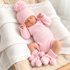 A sleeping newborn baby wearing a pink knit onesie with 'Hello World!' text, a matching pink pom-pom beanie, and pink booties, resting on a white textured blanket