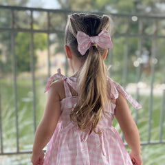Child in a pink gingham dress with a large bow in her hair, standing outdoors.