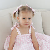 Young girl in a pink gingham dress with bows sitting on a beige couch.