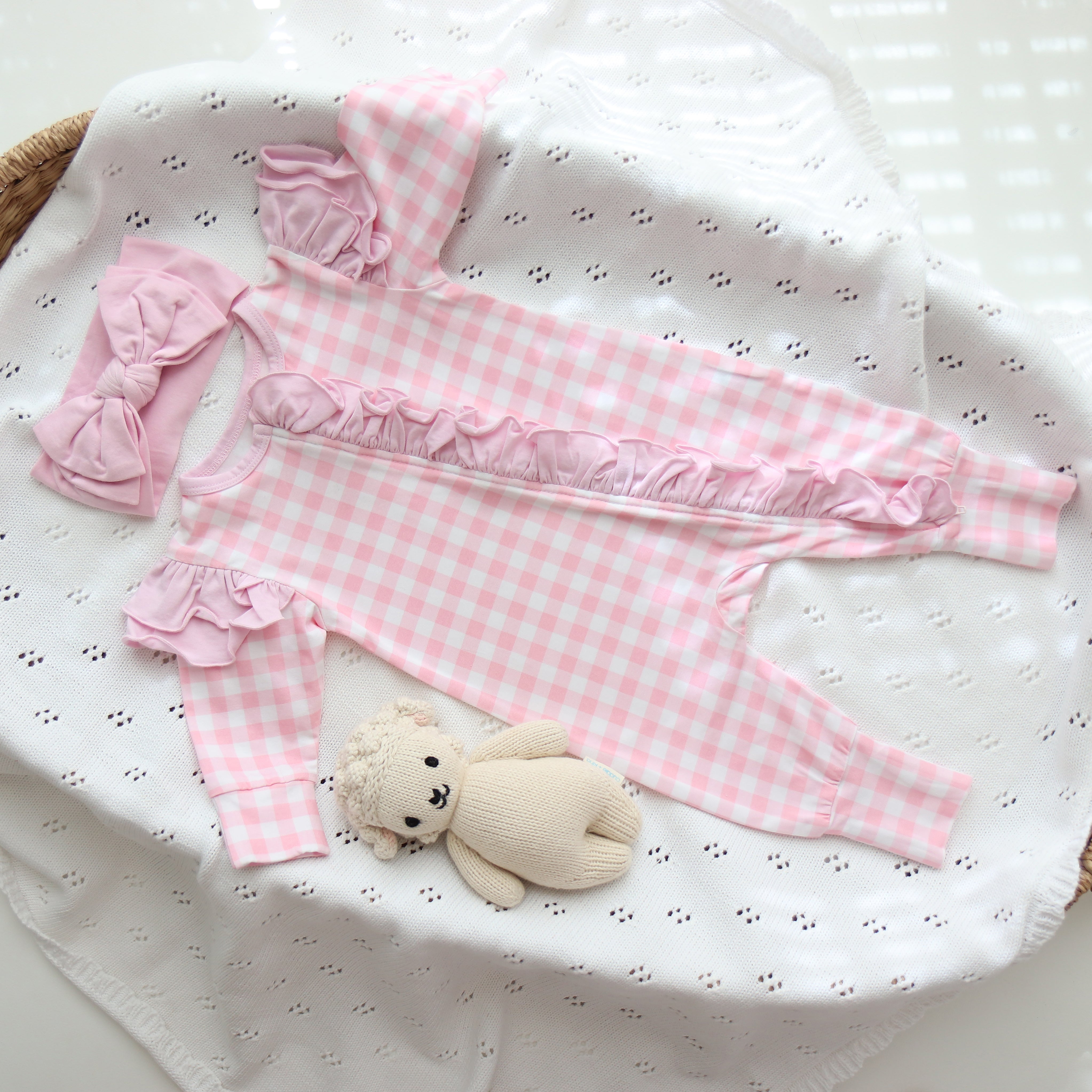 A pink gingham frilly onesie with ruffle frills on the shoulders and fold over cuffs placed on a white fabric, alongside a plush toy.