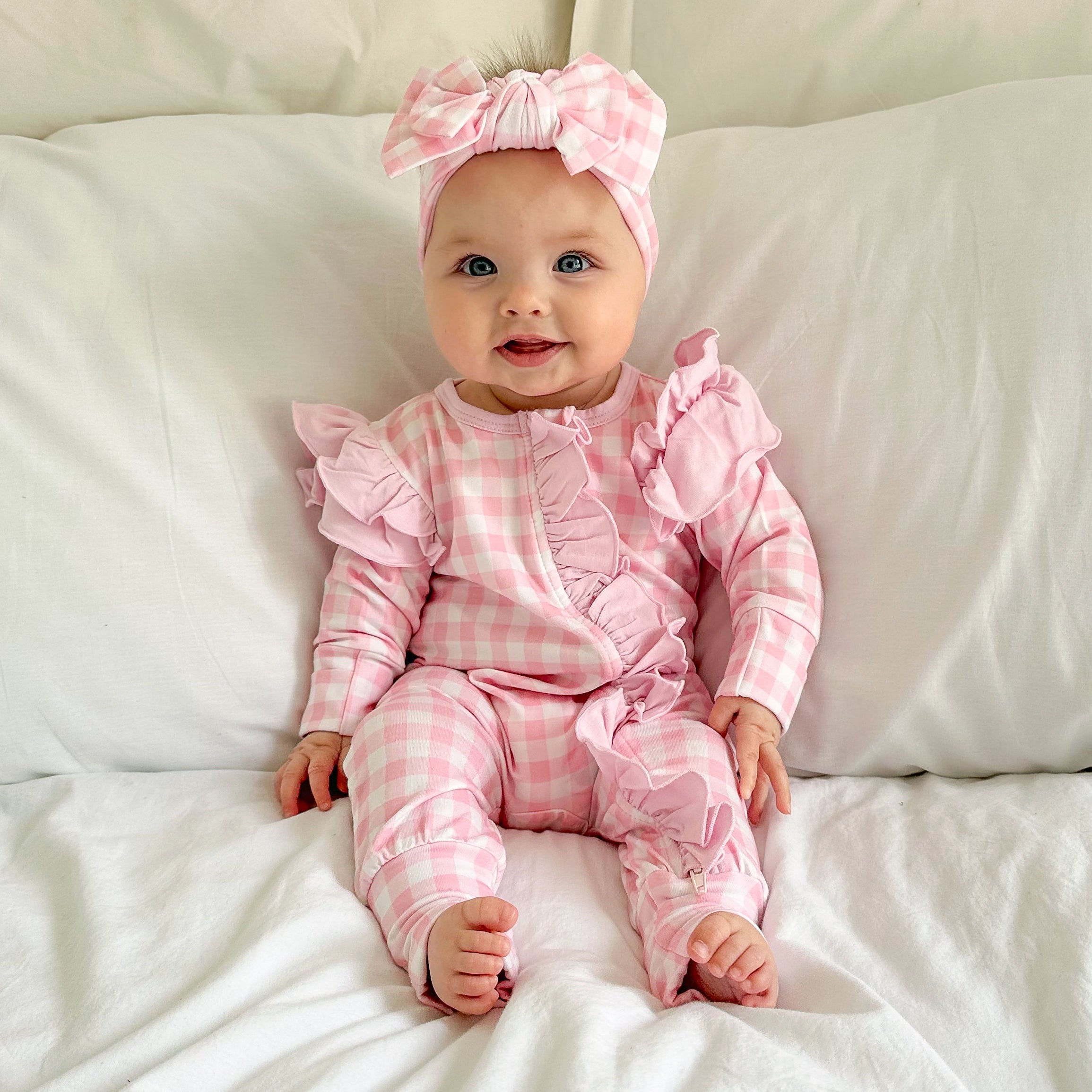 baby dressed in pink gingham frilly onesie with matching one size bow sitting on white blanket