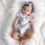Baby wearing a floral playsuit with frill on shoulders and headband on a white blanket
