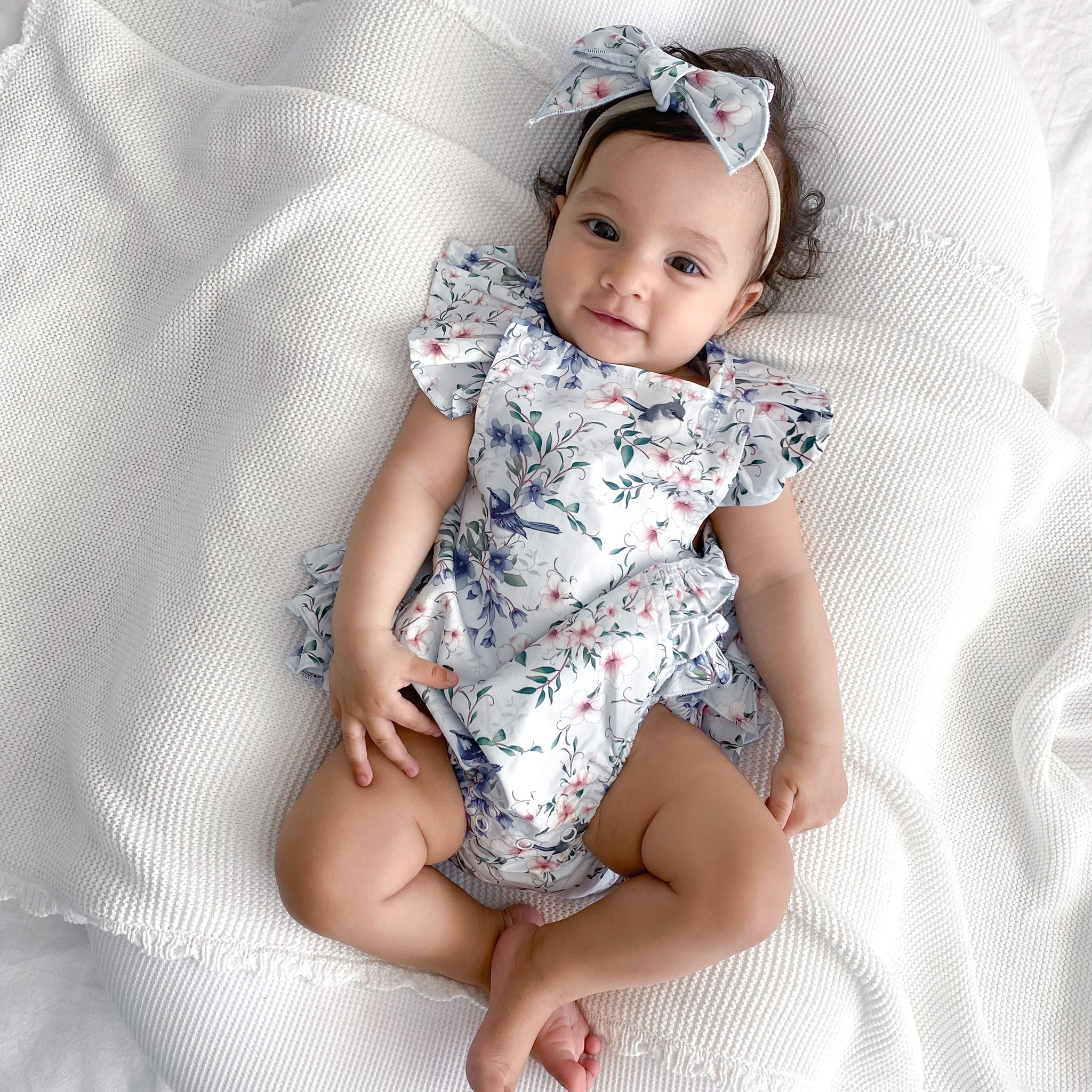 Baby wearing a floral playsuit with frill on shoulders and headband on a white blanket